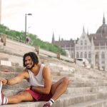 Man with dreadlocks smiling while stretching on city steps near architectural landmark.